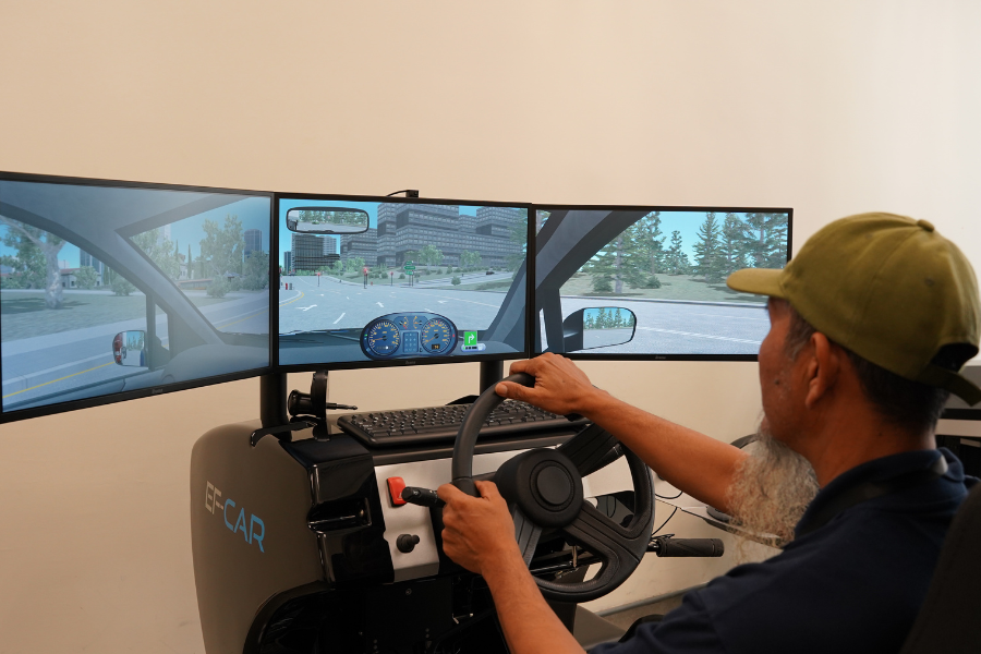 Man using a high-tech driving simulator with a 3-screen urban driving display.